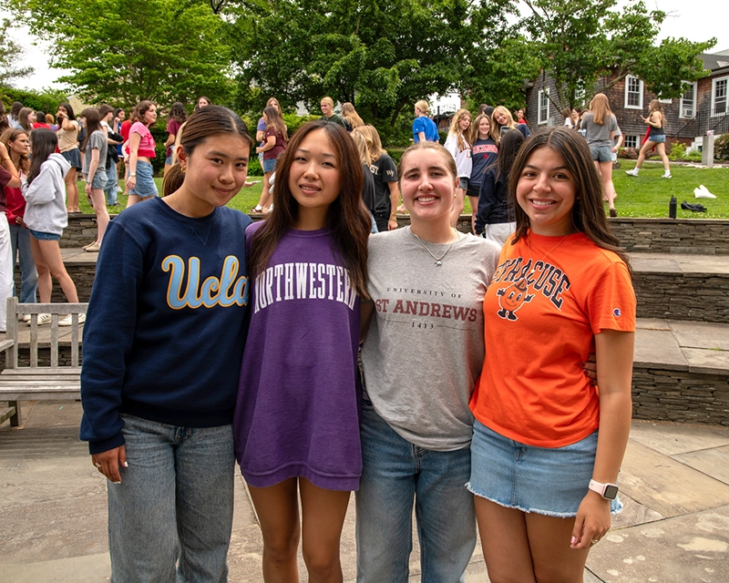 Four young women stand outdoors, smiling at the camera, each wearing a different university shirt: UCLA, Northwestern, St Andrews, and Syracuse. A large group of people and green trees are visible in the background.