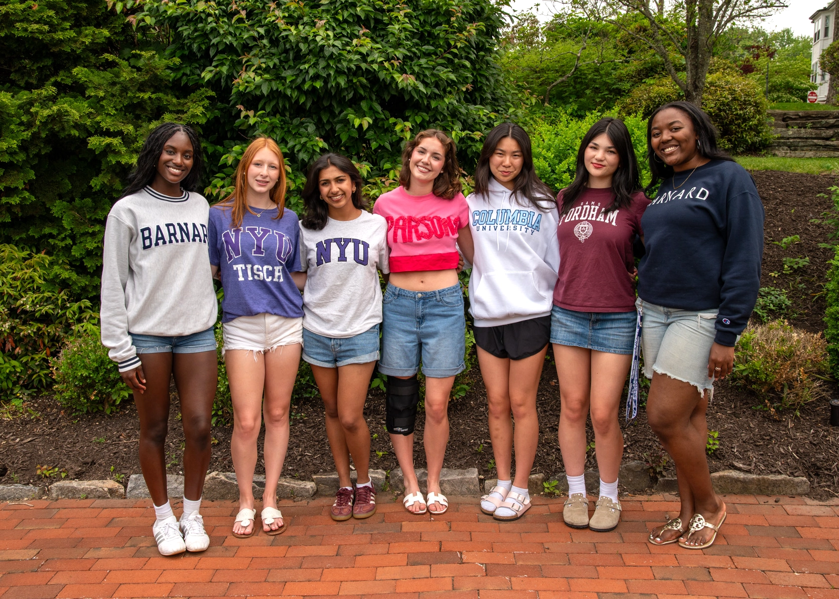 Seven young women stand together outside, smiling. Each wears a college sweatshirt representing different universities, including Barnard, NYU, Harvard, Brown, and Fordham. They stand on a brick path with greenery behind them.