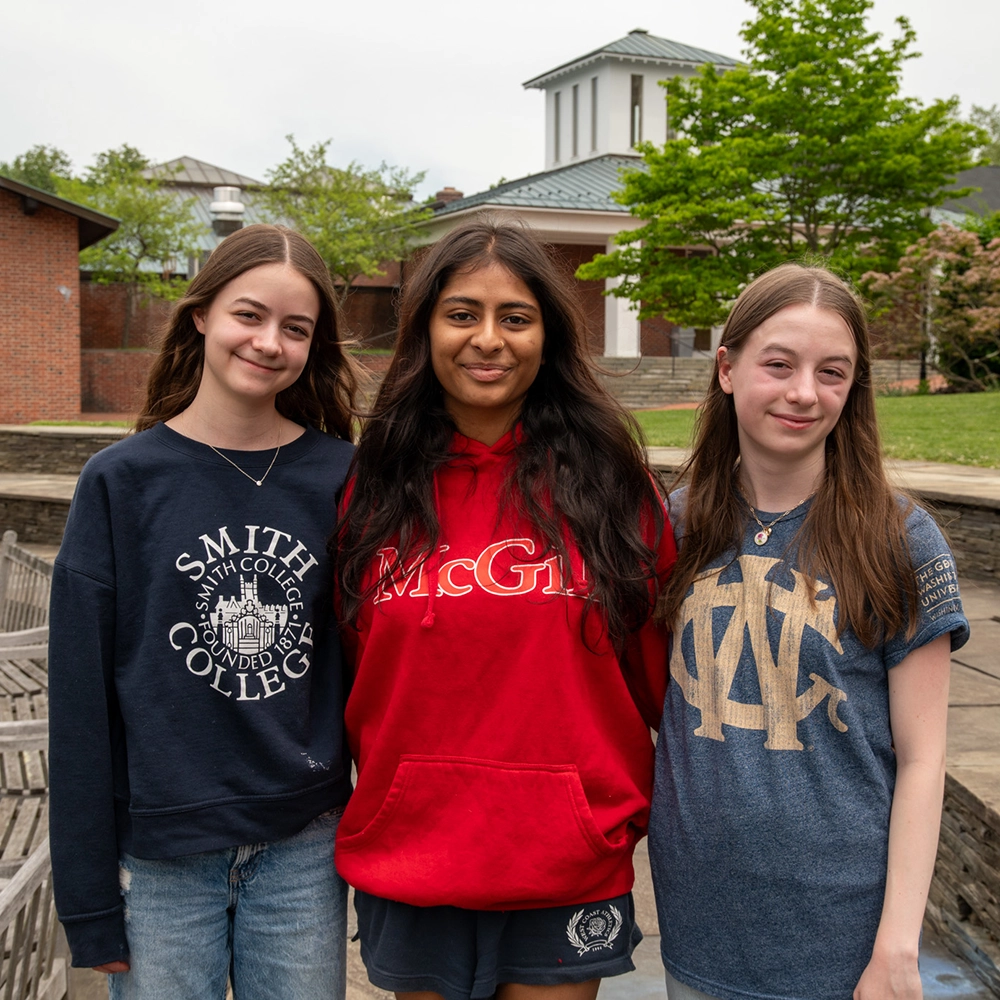 Three young women stand close together outdoors, smiling at the camera. They each wear shirts from different colleges: Smith College, McGill University, and another with a monogram. Trees and buildings are visible in the background.