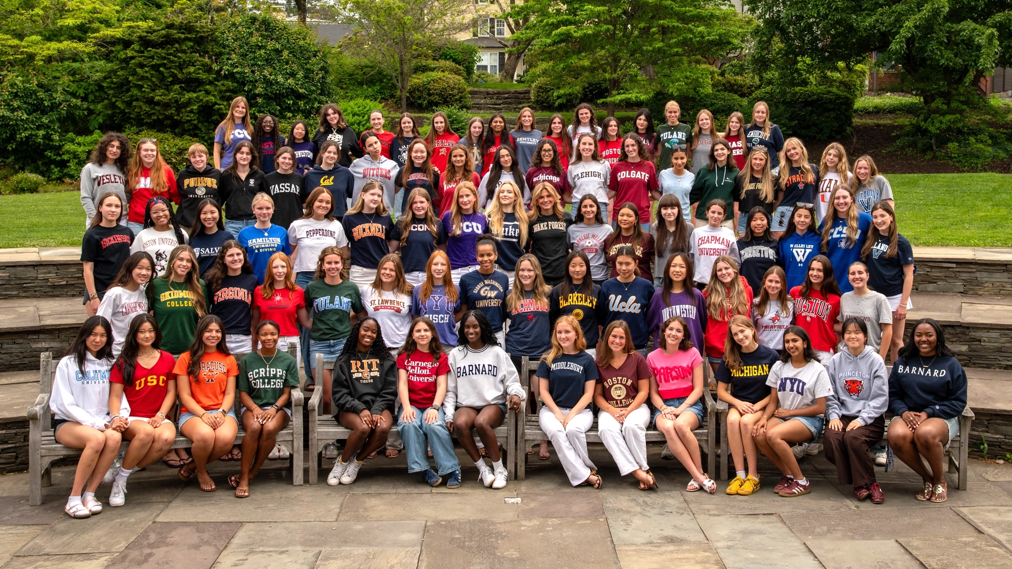 A large group of students sits and stands outdoors, wearing sweatshirts from various colleges and universities. They pose together, smiling, with greenery and stone steps in the background.