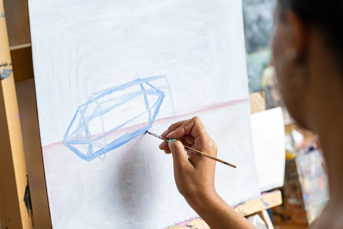 A person paints a geometric blue shape on a blank canvas using a paintbrush. Only their hand and part of their head are visible. The scene appears to be in an art studio.