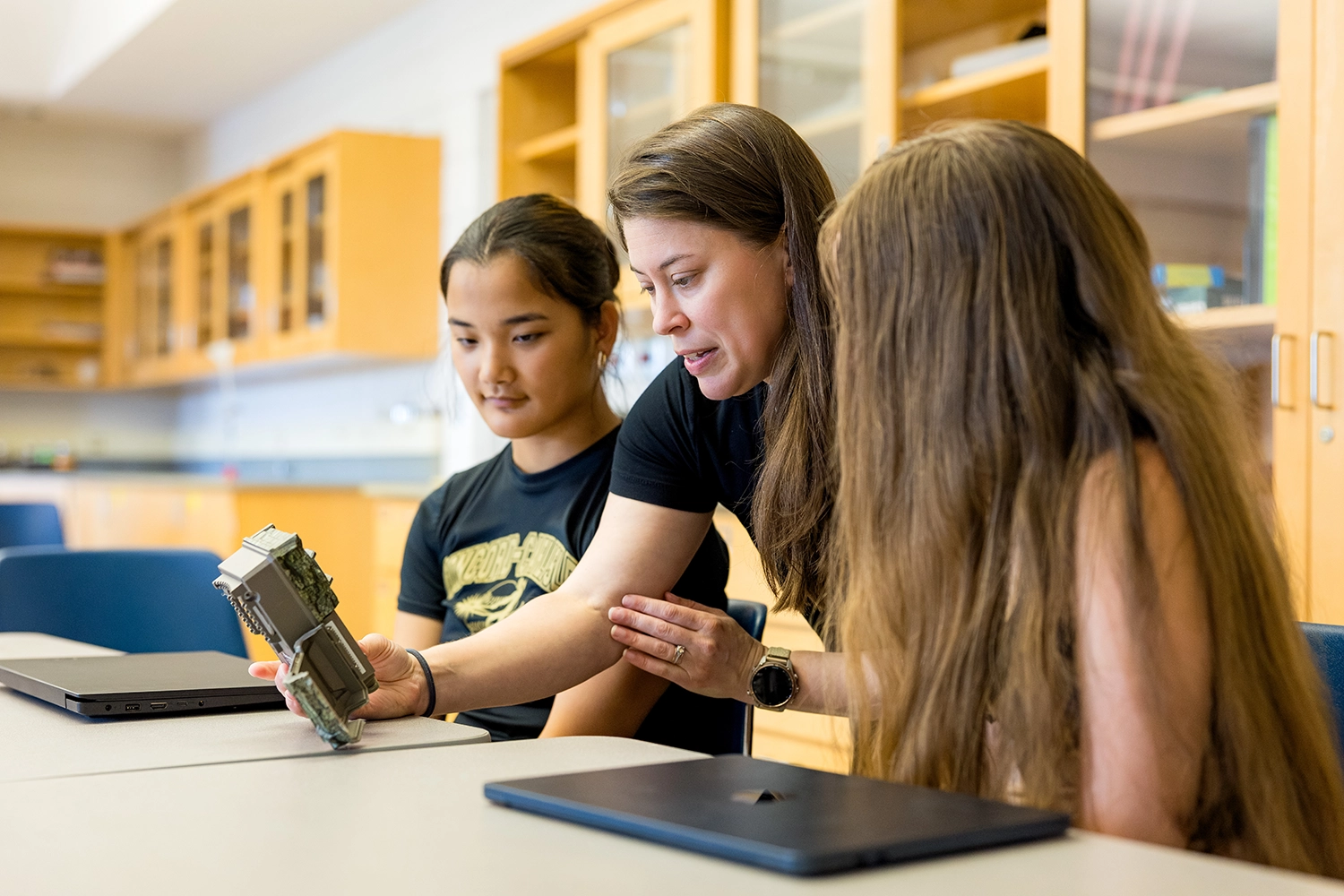 A teacher shows two students a computer component at a classroom table. The students watch attentively, and laptops are on the table in front of them. Shelves with books and supplies are visible in the background.