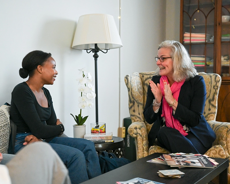 Two women sit and smile while talking in a cozy living room. One gestures enthusiastically with her hands, and the other listens attentively. Magazines and books are on the table in front of them.