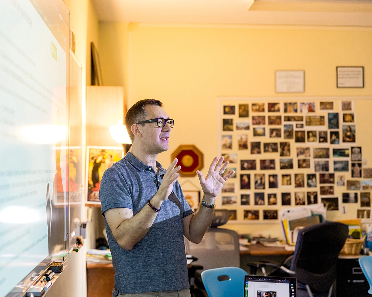 A man wearing glasses gestures while speaking in a classroom with a whiteboard, photos on the wall, and desks with computers and chairs in the background.
