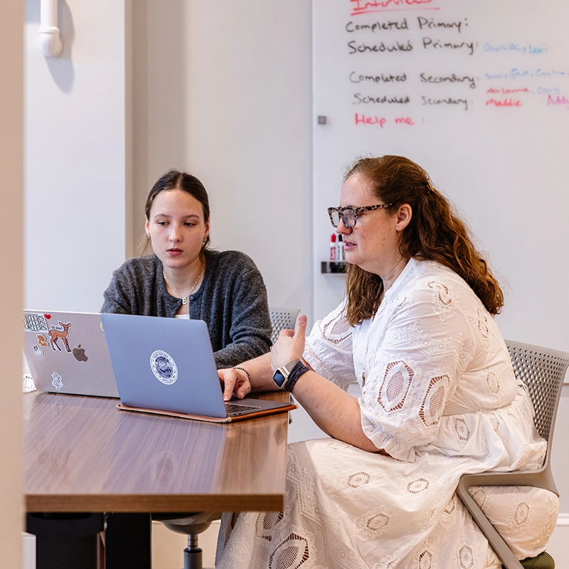 Two women sit at a table working on laptops in an office. One gestures while the other listens. A whiteboard with colorful writing hangs in the background.