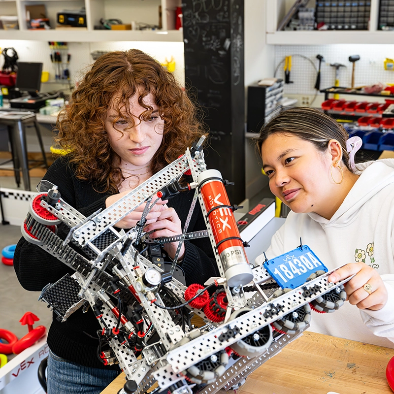 Two students work together on assembling a robot in a classroom filled with tools and parts, focusing intently on adjusting its mechanisms.