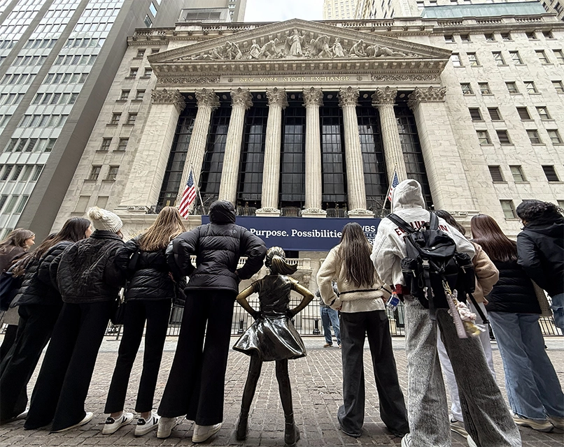 A group of people stand with their backs to the camera, facing the Fearless Girl statue and the New York Stock Exchange building. Two American flags hang above the entrance.