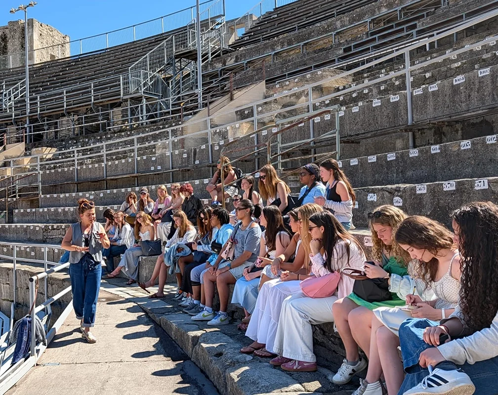 A group of people sits on stone stadium steps under sunlight, listening to a guide. The seats are numbered, and the group appears relaxed, some taking notes or holding bags. The stadium is mostly empty.