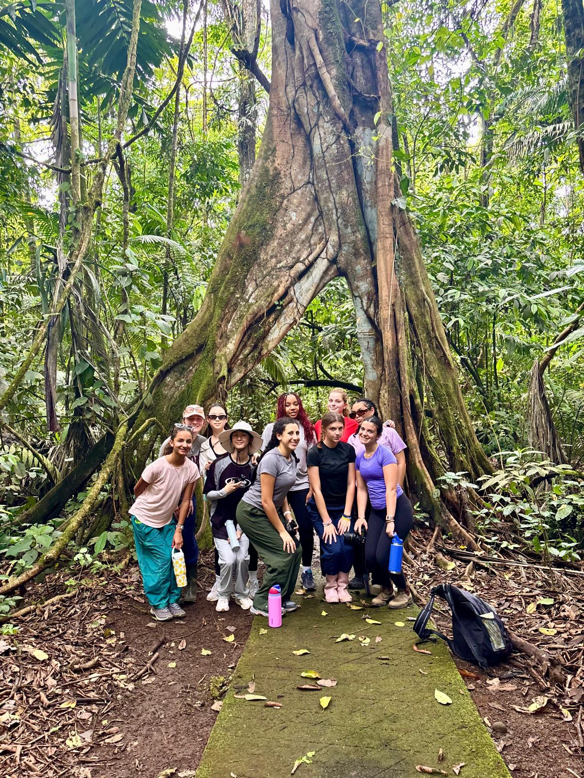 A group of people pose in front of a large tree with exposed roots in a lush, green forest. Some carry water bottles and wear hats, and a backpack rests on the ground nearby.