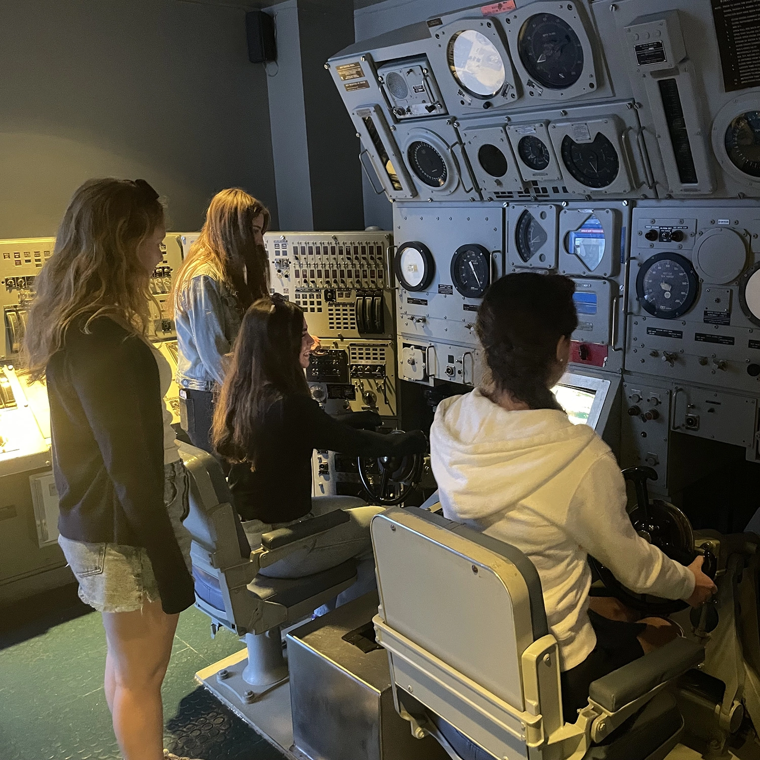 Four young women interact with a large control panel filled with dials and screens in a dimly lit room. Two are seated at the controls, while two stand behind watching closely.