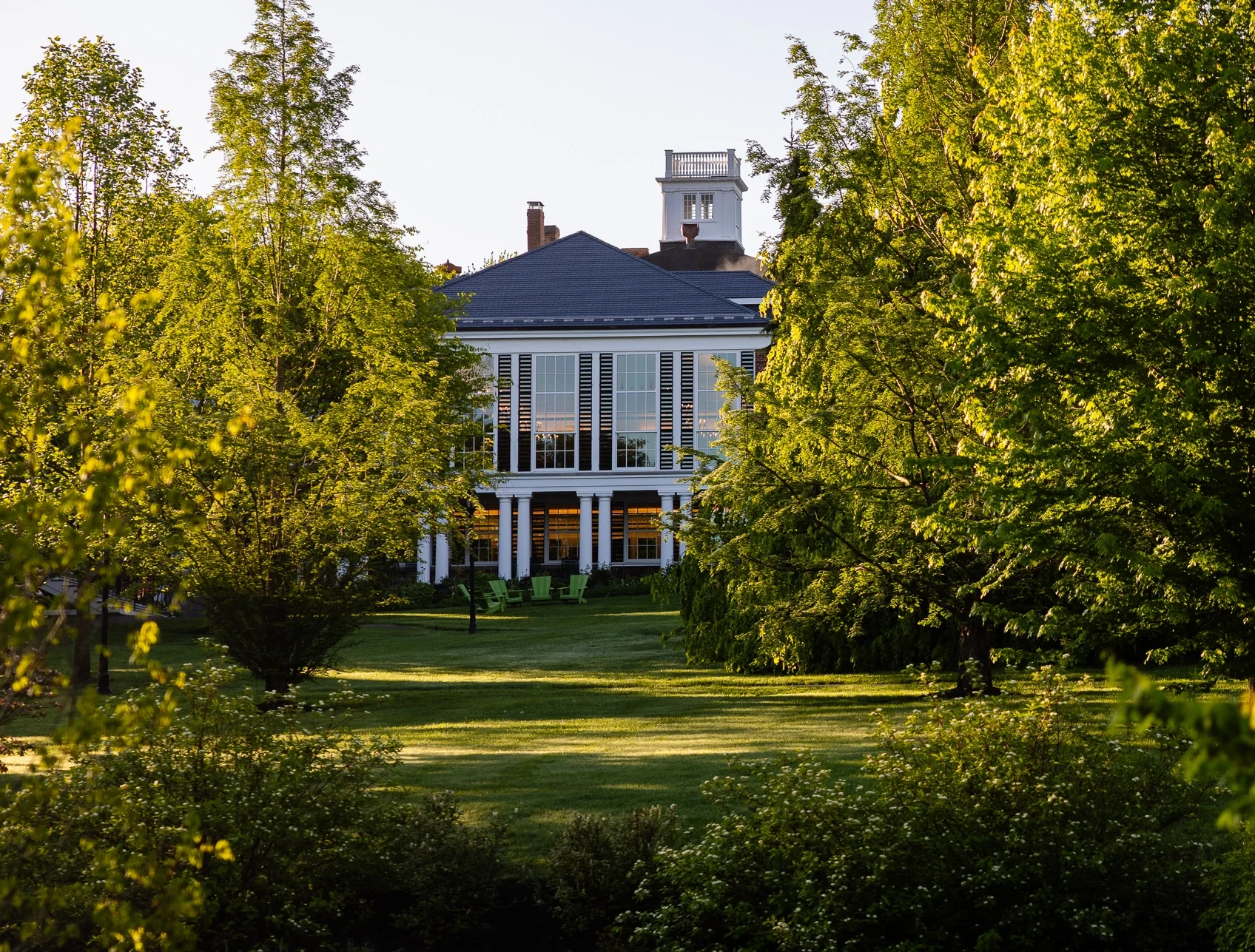 A large, white, two-story house with tall columns is partially hidden by lush green trees and grass in bright daylight.