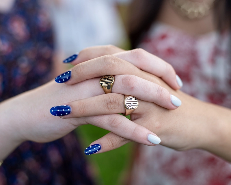 Two people with manicured nails—one with blue and white polka dots, the other with light blue—hold hands. Both wear gold rings with the MPS signet. Their floral dresses are visible in the background.