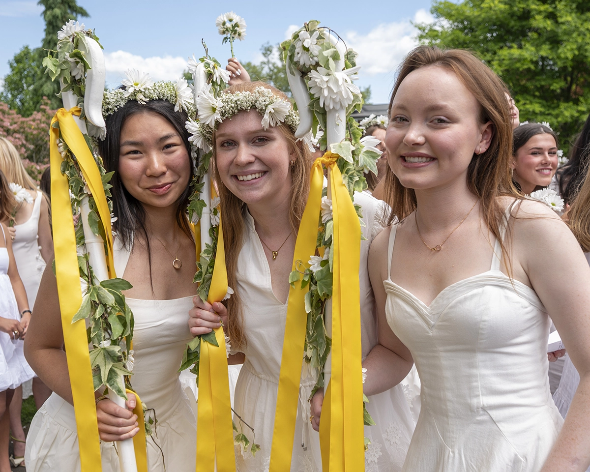 Three young women in white dresses and flower crowns smile and hold decorated poles with yellow ribbons at an outdoor celebration; others in white are in the background, trees and blue sky visible.