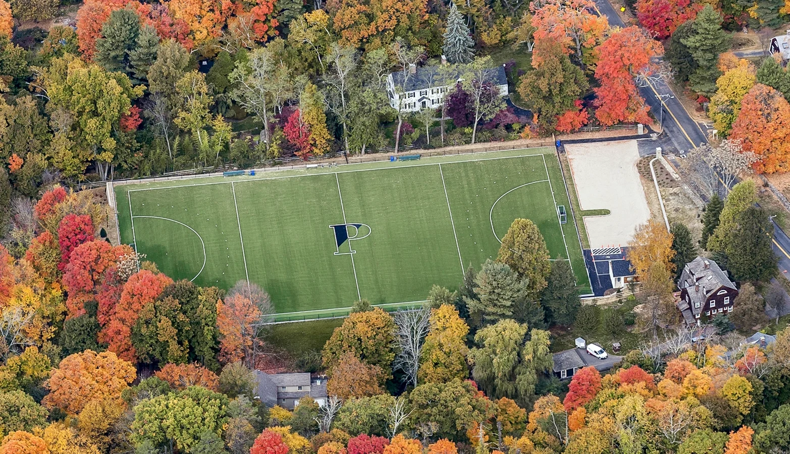 Aerial view of a green sports field with a large “P” at the center, surrounded by trees with autumn foliage in red, orange, and yellow hues, with a few houses nearby.