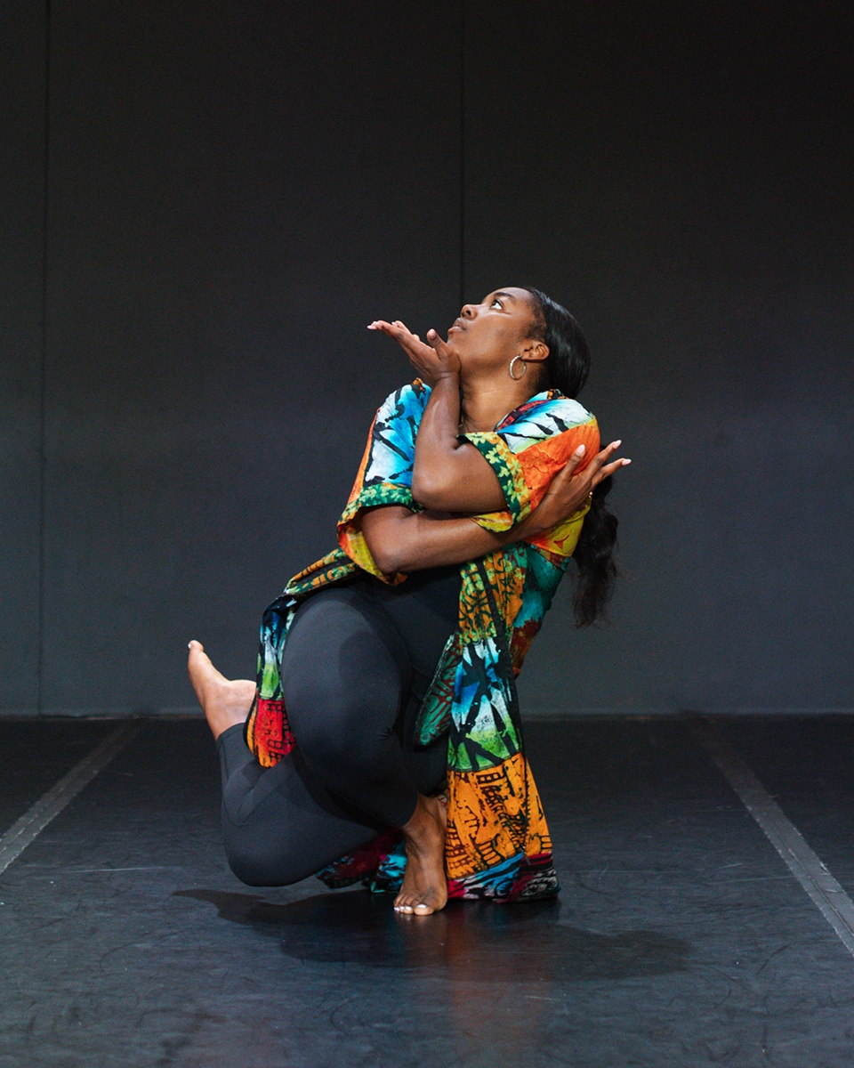 A woman in colorful clothing performs a modern dance pose on stage, balancing on one leg with arms crossed and looking upward, against a plain dark background.