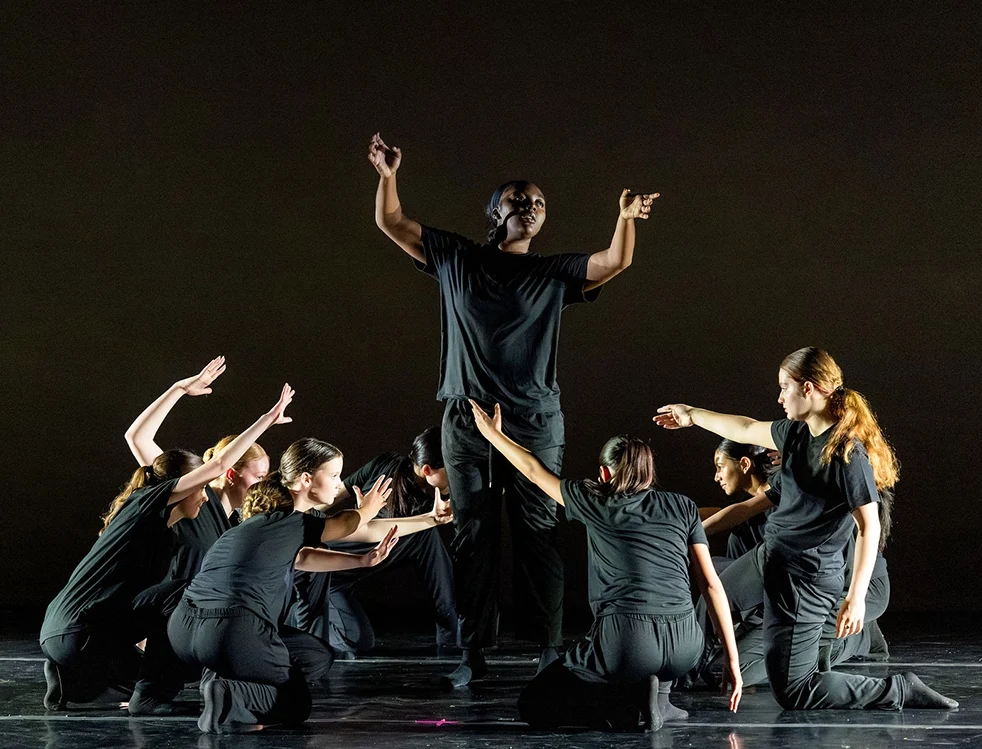 A group of dancers in black outfits kneel and extend their arms toward a central standing dancer, who raises their hands against a dark background.