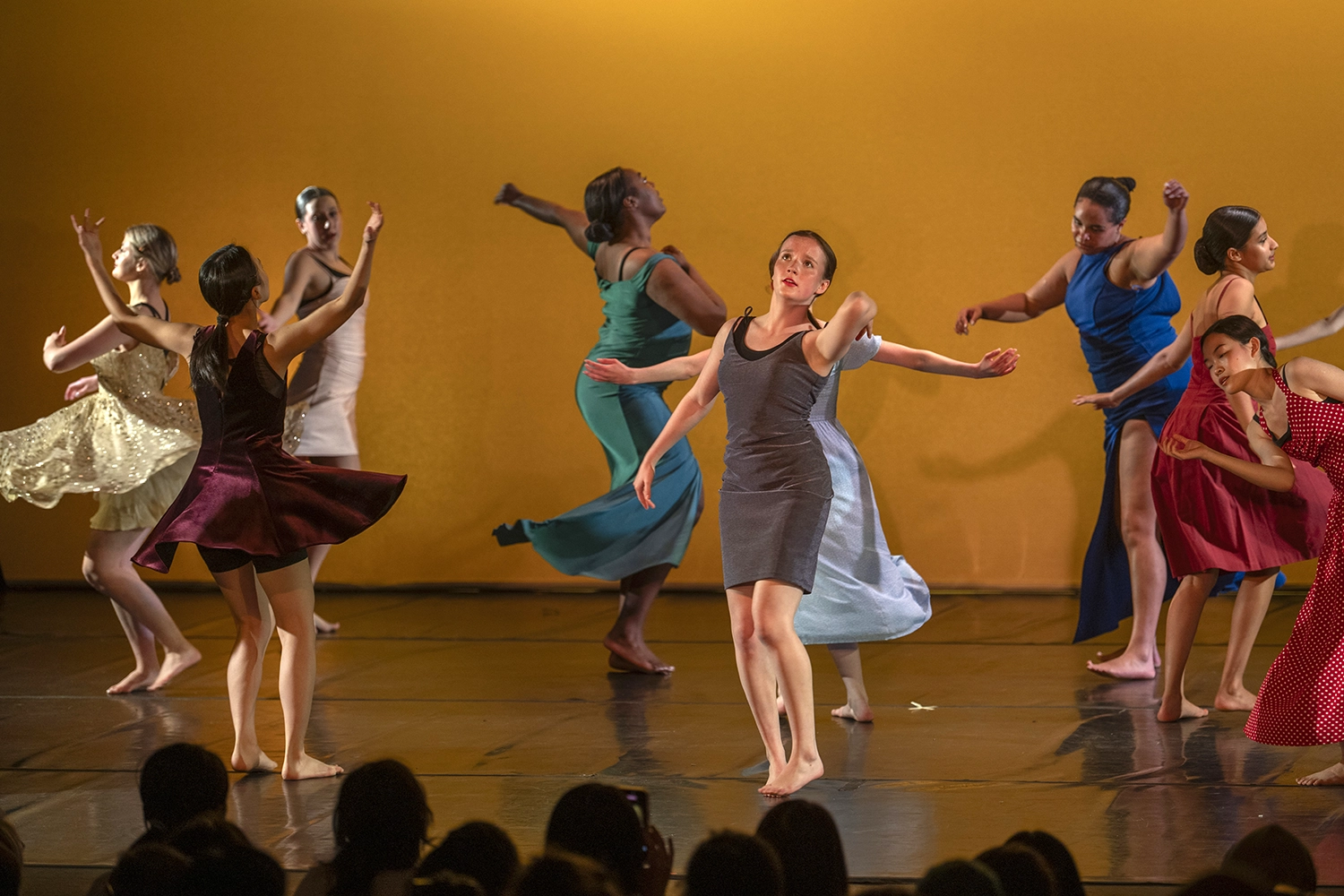 A group of dancers in colorful dresses perform barefoot on stage against a warm yellow backdrop, striking expressive poses as an audience watches.