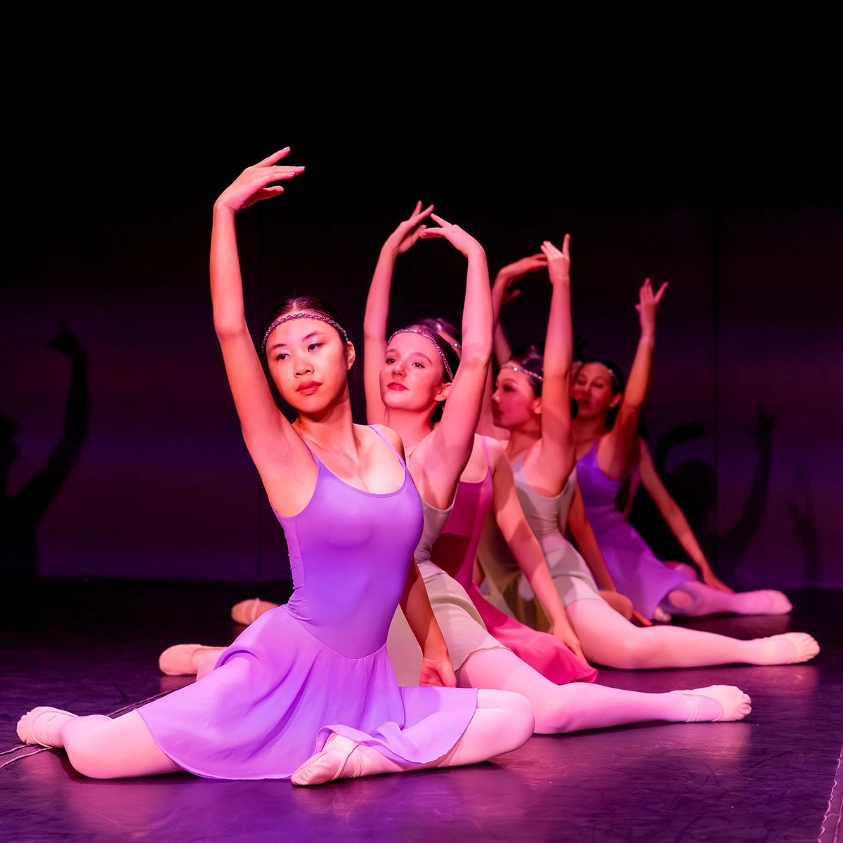 Four ballet dancers in colorful leotards perform a seated pose on stage, each with one arm gracefully raised, under pink and purple stage lighting.