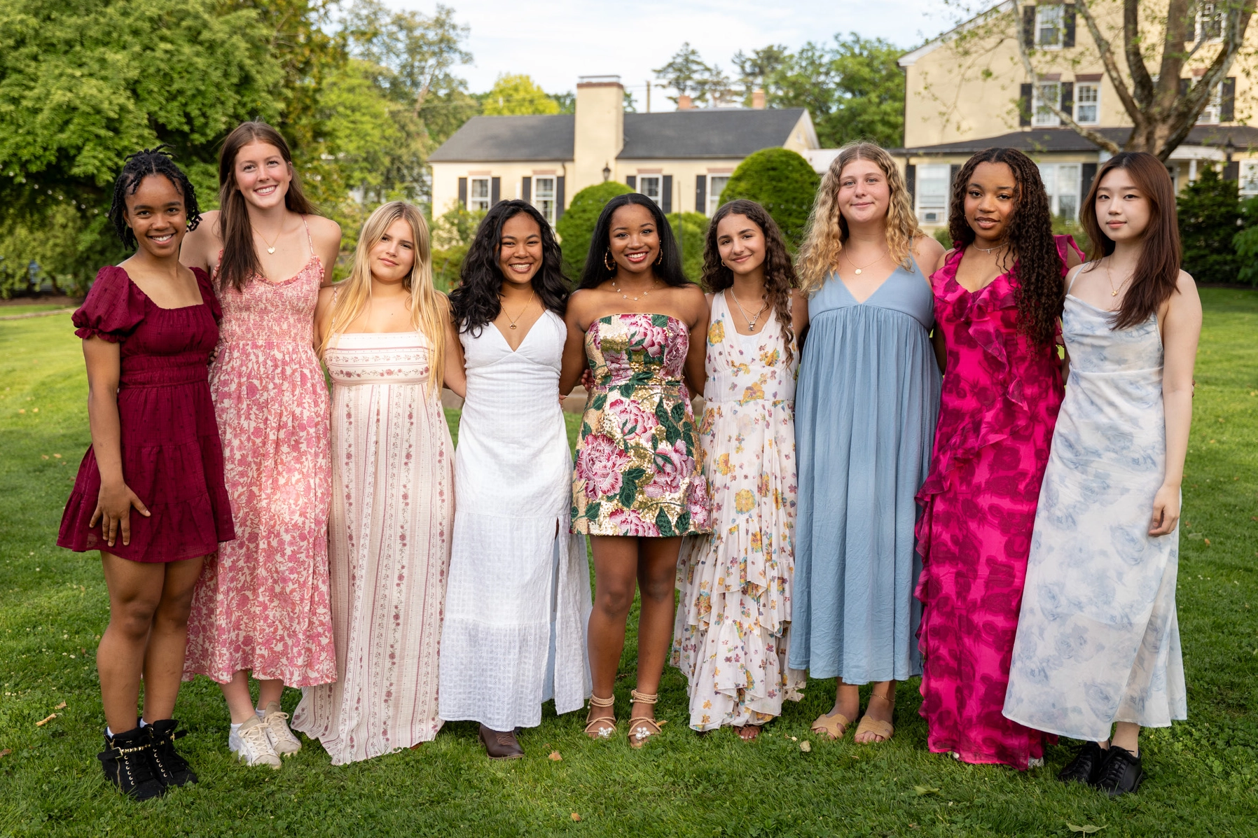 A group of eight young women stand together outside on grass, smiling and dressed in colorful summer dresses. Trees and houses are visible in the background on a sunny day.