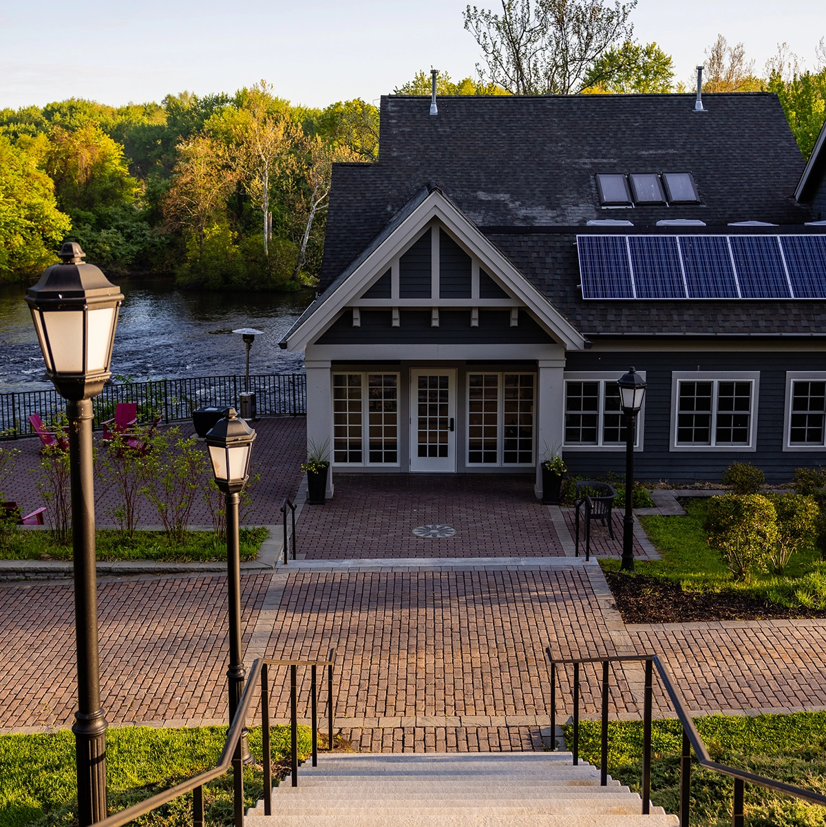 A building with solar panels on the roof sits by a river, surrounded by trees. Brick steps and lamp posts lead down to the entrance, and sunlight casts long shadows on the patio.