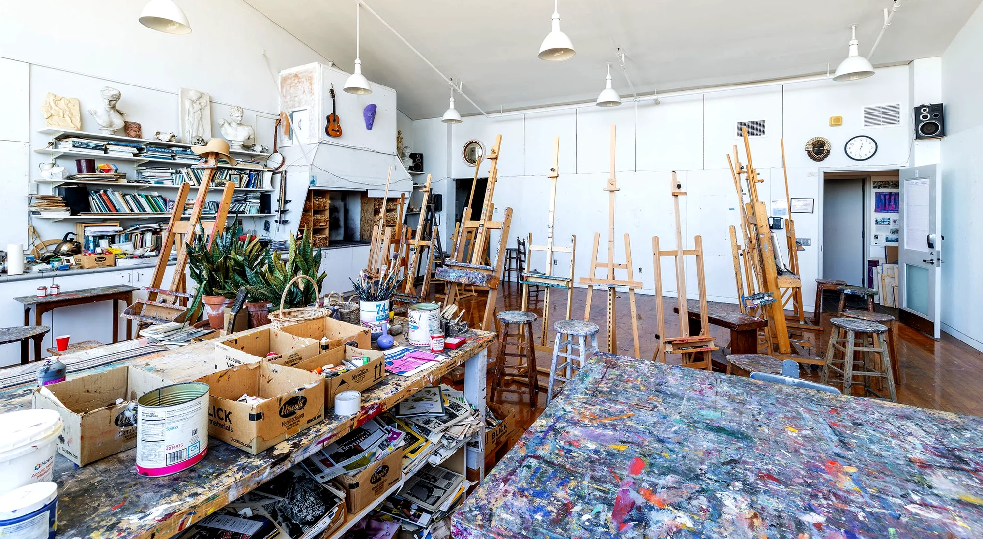 Bright art studio with easels, stools, and tables covered in paint. Shelves with art supplies, plants, and books line the wall. Paint cans and brushes are scattered around. White walls and high ceiling with hanging lights.
