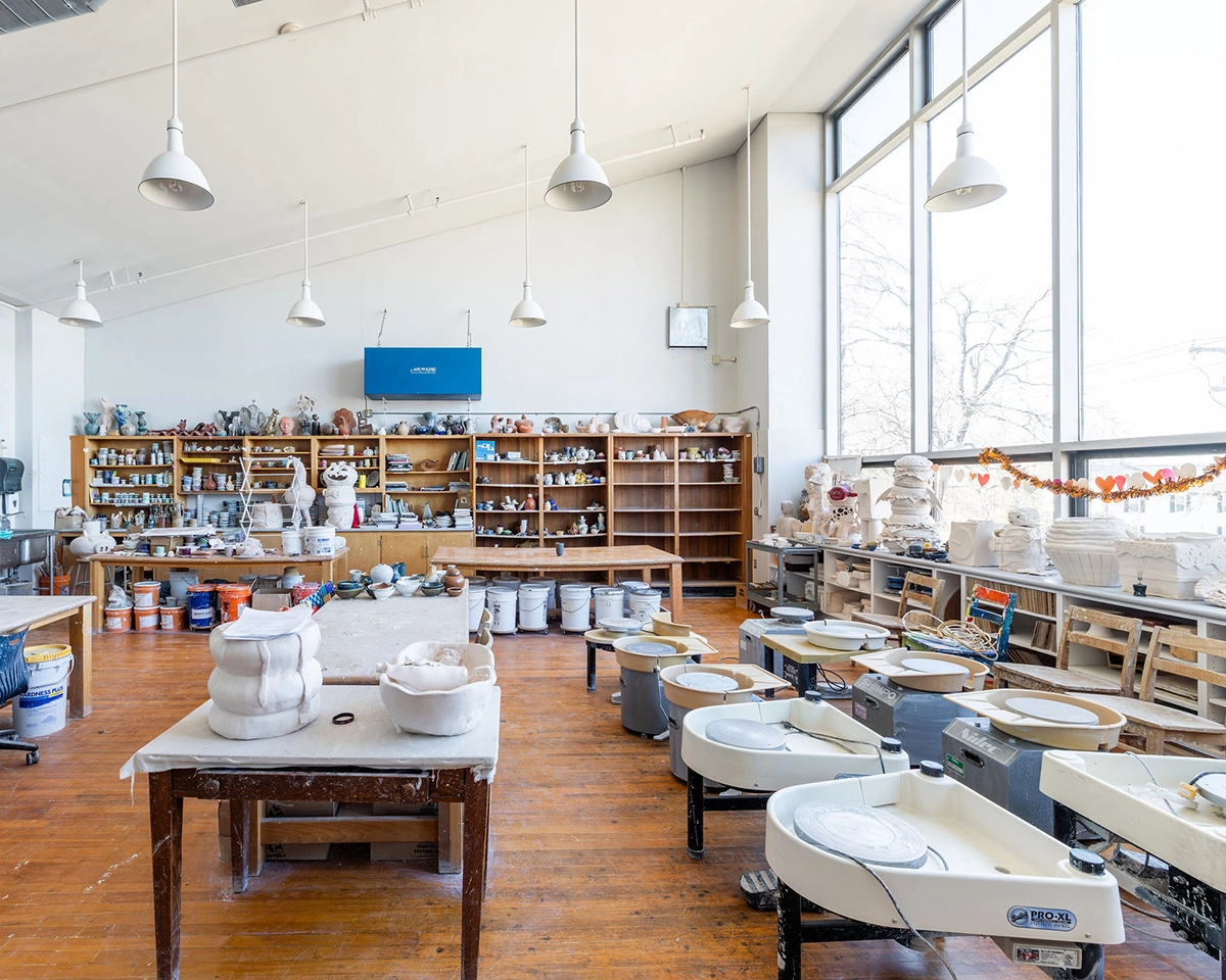 A bright ceramics studio with pottery wheels, a table with clay sculptures, shelves filled with pottery supplies, and large windows letting in natural light.