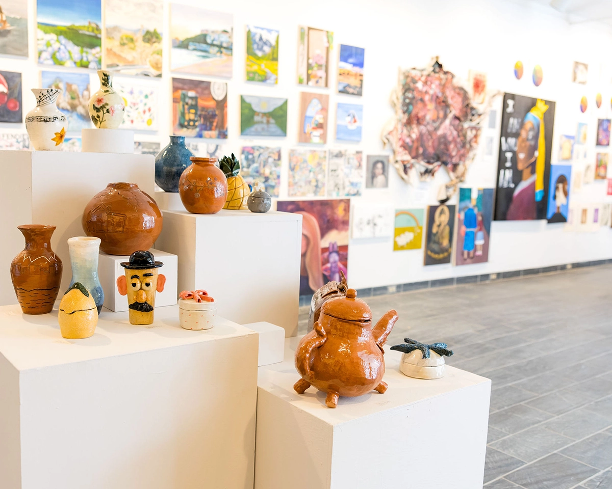 A gallery displays colorful paintings on white walls and showcases various handmade ceramic pots, vases, and figures on white pedestals in a bright, modern space.
