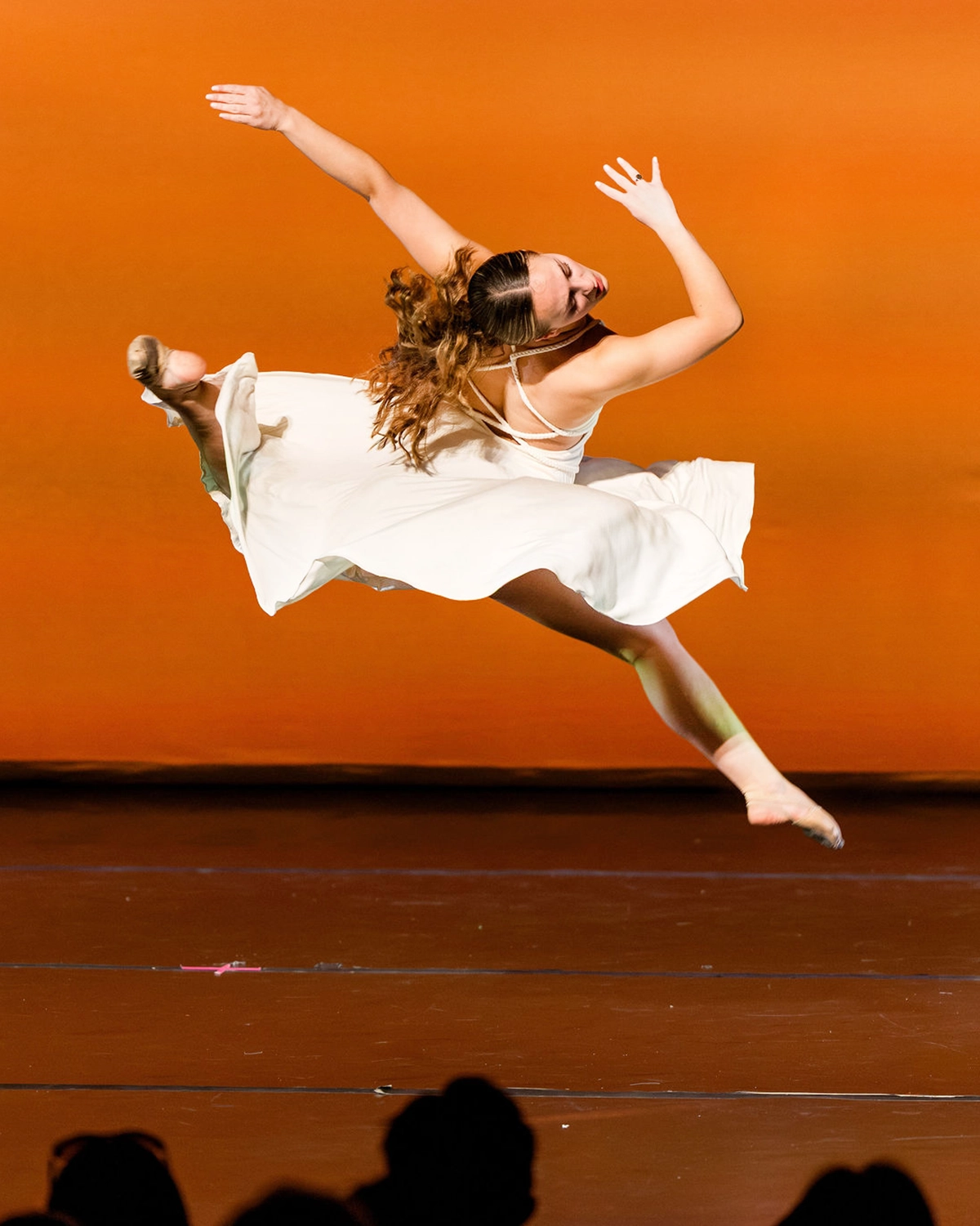 A dancer in a white dress leaps gracefully across a stage with an orange background, her hair flowing and arms extended, capturing a dynamic moment in mid-air.