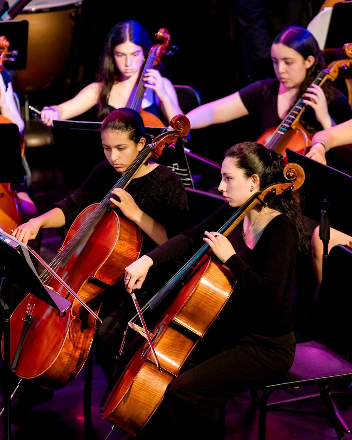 Four young women play cellos in an orchestra, seated closely together on stage, wearing black outfits and focused on their music during a performance.