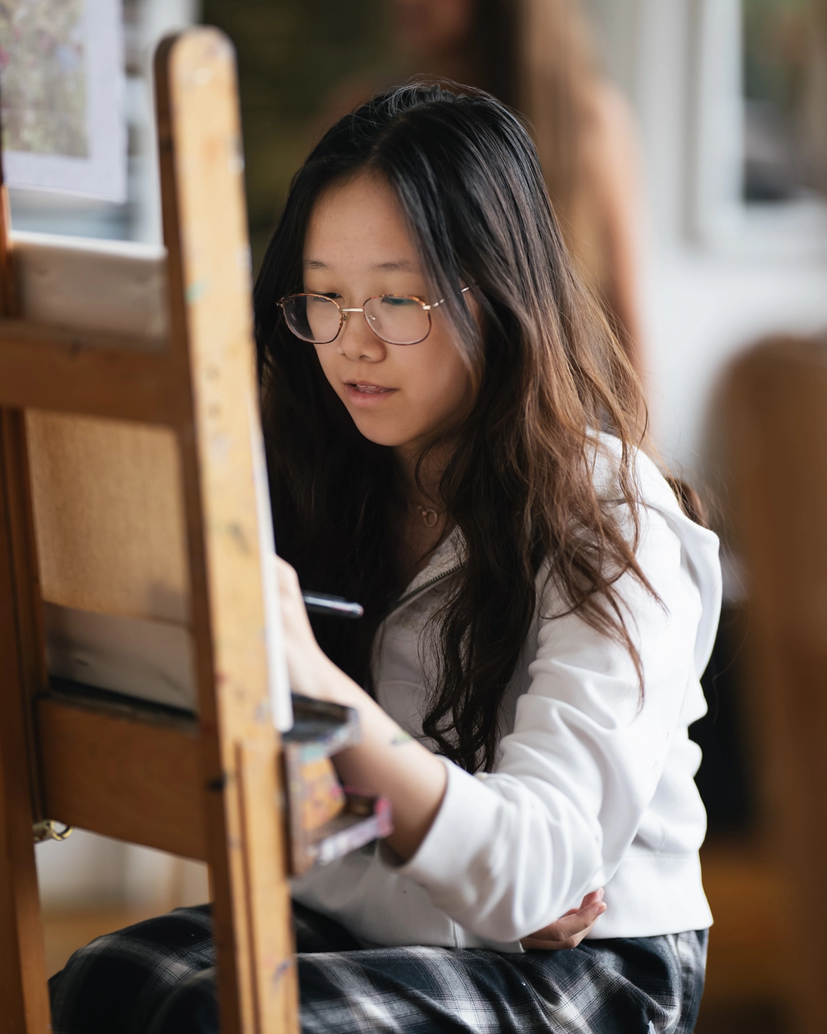 A young woman with long hair and glasses draws at an easel, wearing a white top and plaid pants, focused on her artwork in a bright, indoor studio.