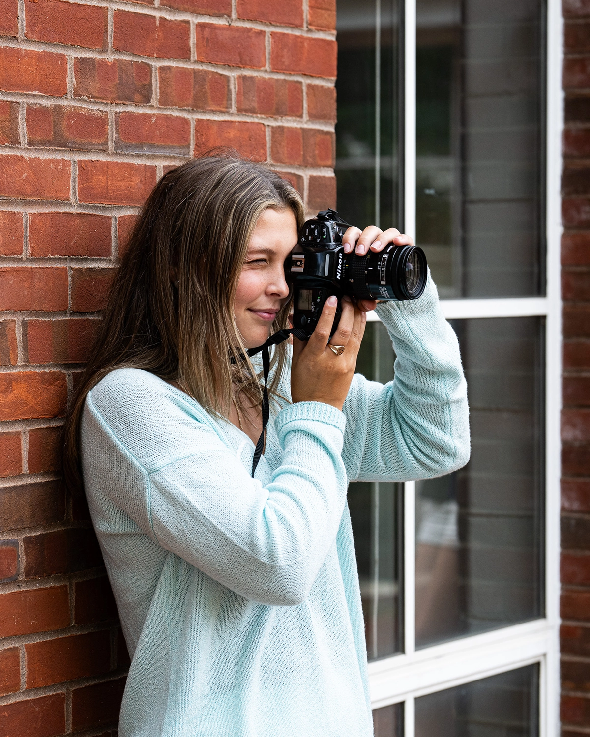 A woman with long hair wearing a light blue sweater stands by a brick wall and a window, holding a camera up to her face as she takes a photograph.