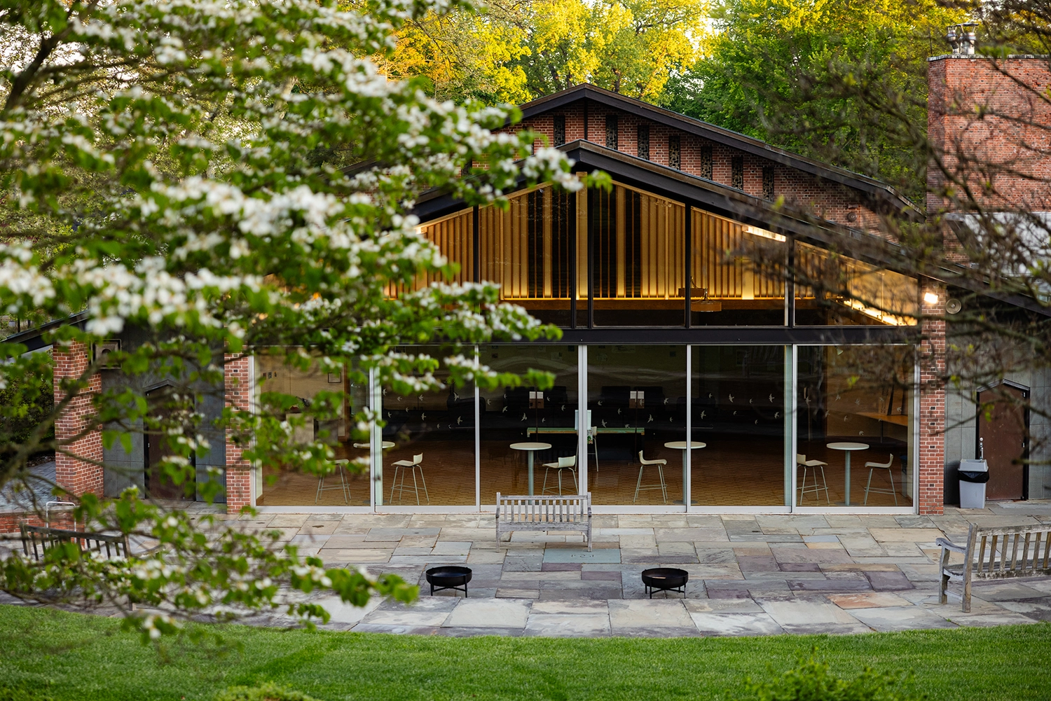 A modern building with large glass windows and brick walls sits behind a patio with benches and tables, surrounded by green grass and leafy trees in spring.