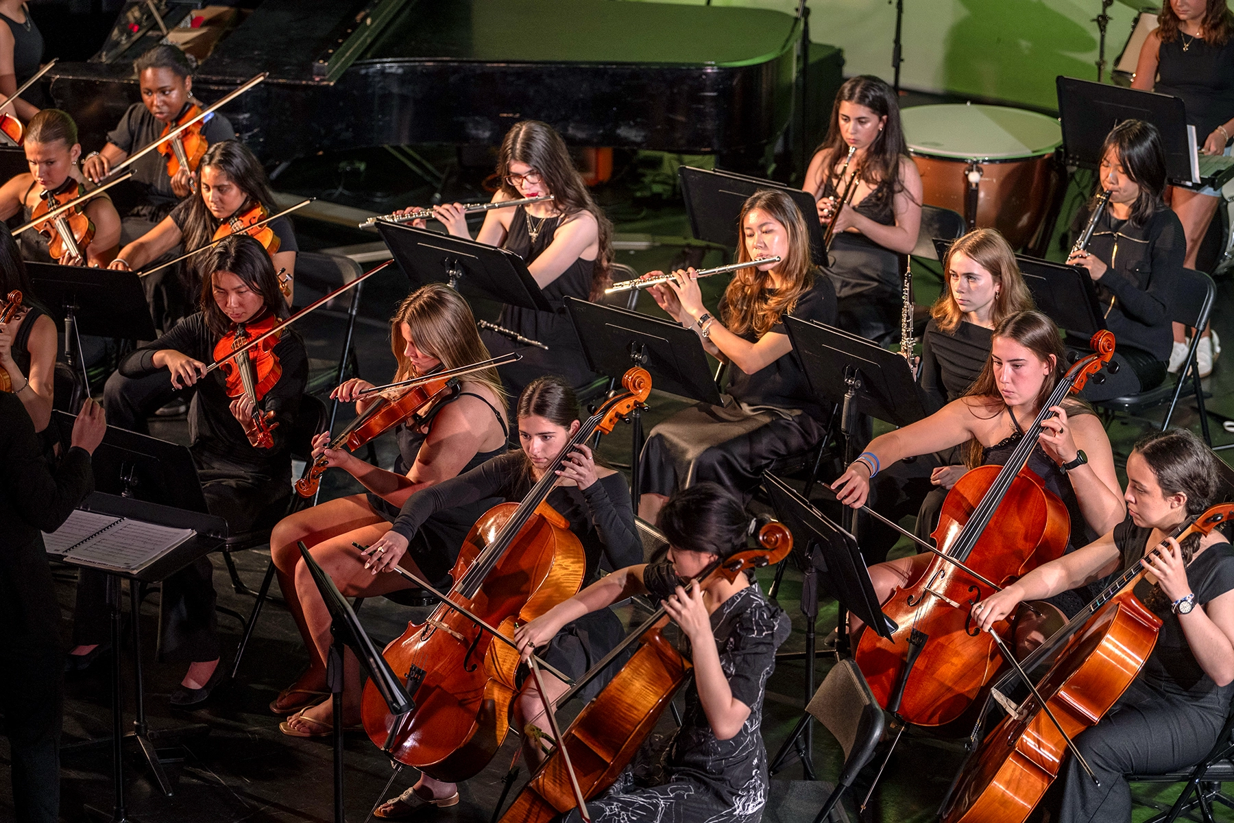 A group of musicians, mainly women, play string and wind instruments in an orchestra. They are seated in rows, dressed in black, and are focused on their sheet music during a performance.