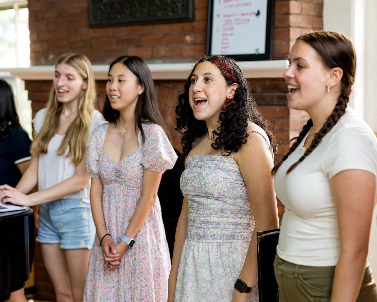 Four young women stand side by side, singing indoors. They appear cheerful and focused, wearing summer clothing. A piano and brick wall are visible in the background.