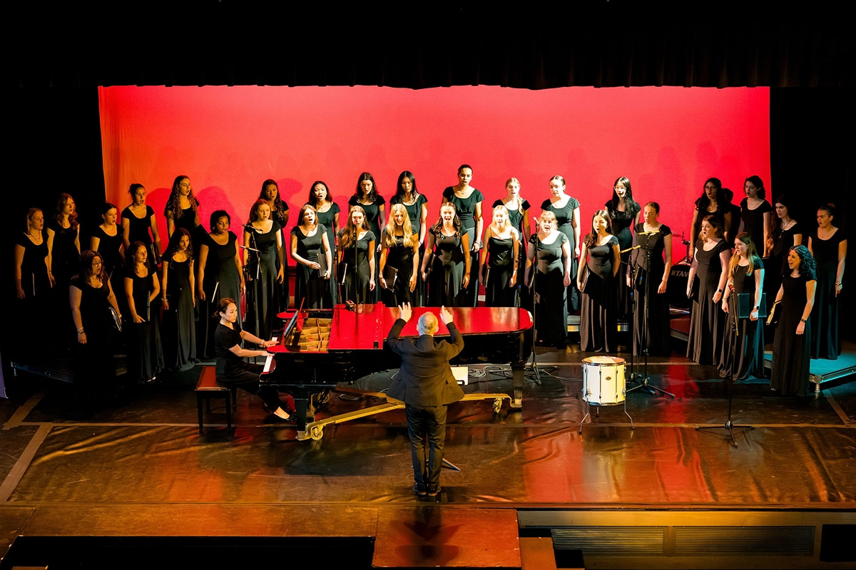 A choir of women in black dresses sings on stage, led by a conductor in front of a grand piano, with a red backdrop.