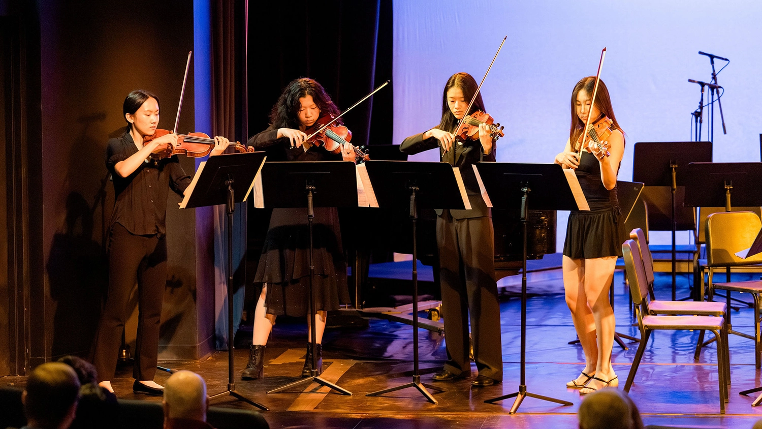 Four women playing violins on stage, dressed in black, standing behind music stands. Stage lights illuminate them, with an empty chair and microphone stand visible in the background.