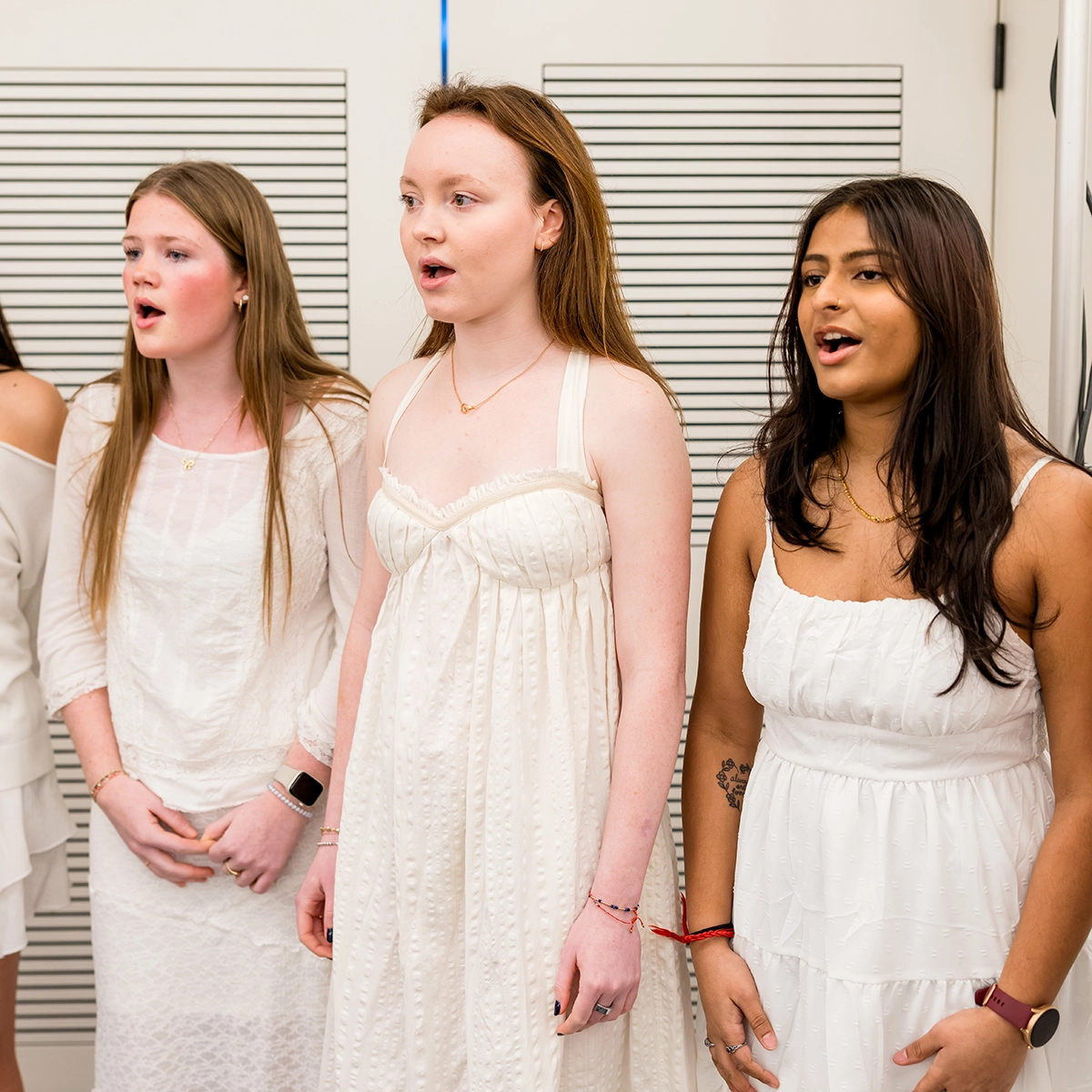 Three young women in white dresses are standing together singing. They appear engaged and focused, possibly performing in a choir or musical event, against a bright indoor background.