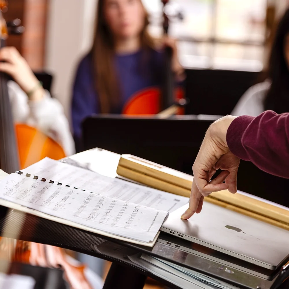 A person points at a laptop on a music stand with sheet music. Several blurred people in the background appear to hold musical instruments, possibly in a rehearsal setting.