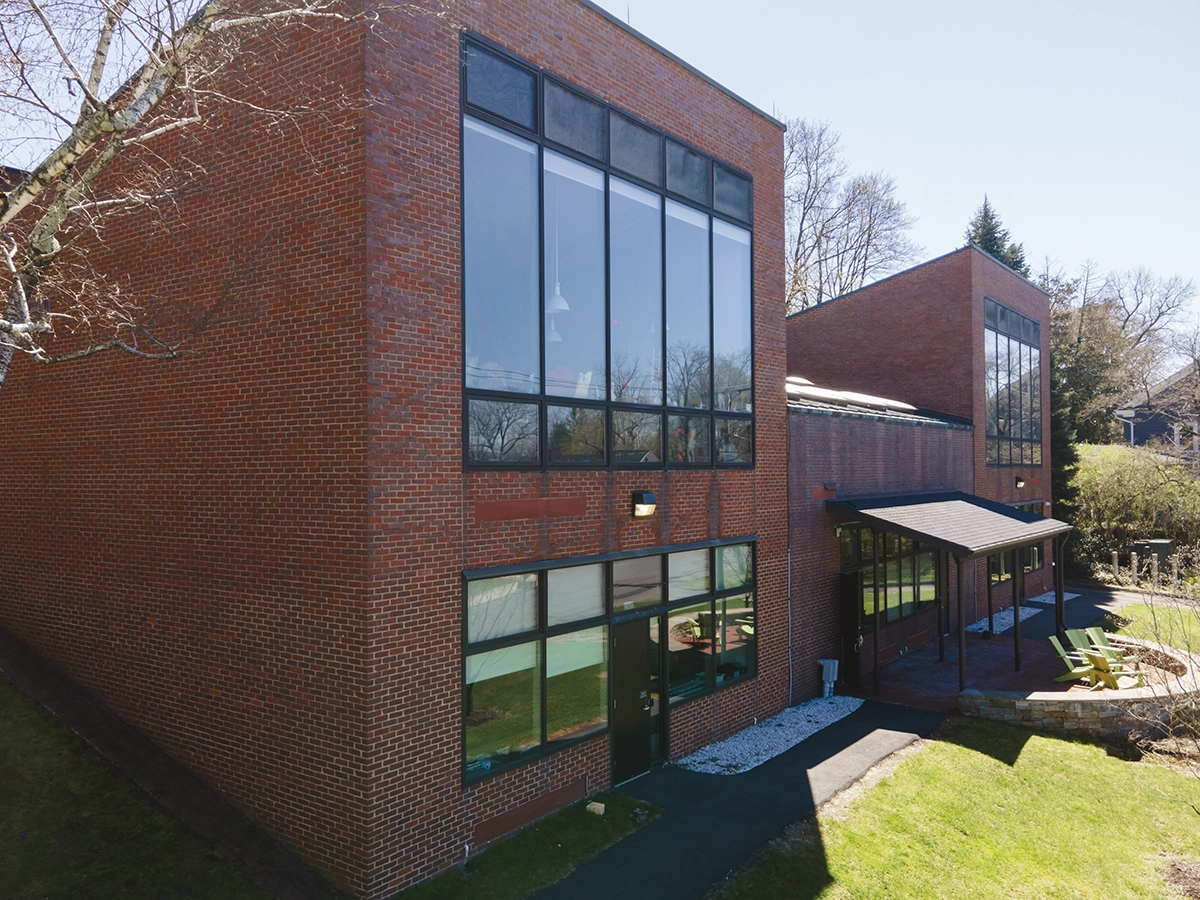 Modern red brick building with large tall windows, a sloped roof, and a small covered entryway. The structure is surrounded by grass, trees, and a stone-lined path on a sunny day.