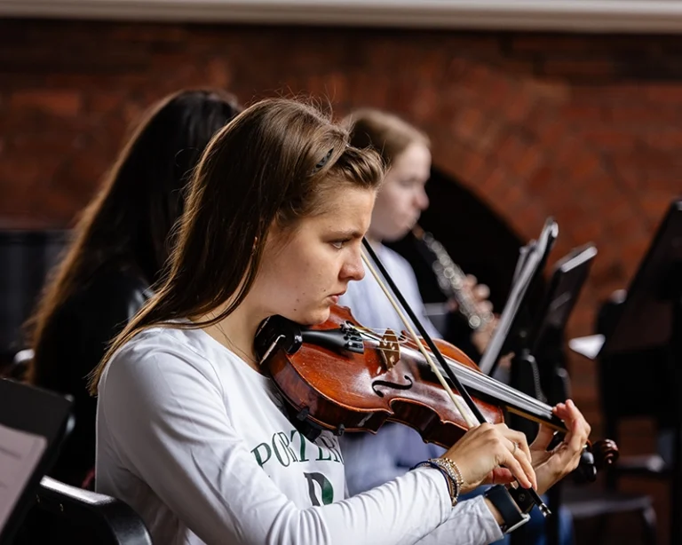 A young woman with light brown hair plays the violin in an indoor setting, with two other musicians blurred in the background. A brick wall is visible behind them.