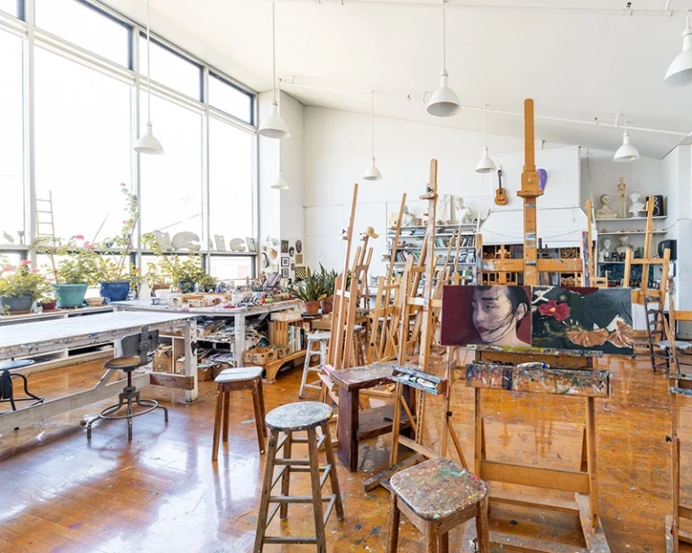 Bright art studio with large windows, wooden floors, easels, stools, and art supplies. A portrait painting of a woman is on one easel. Plants and shelves with materials line the walls.