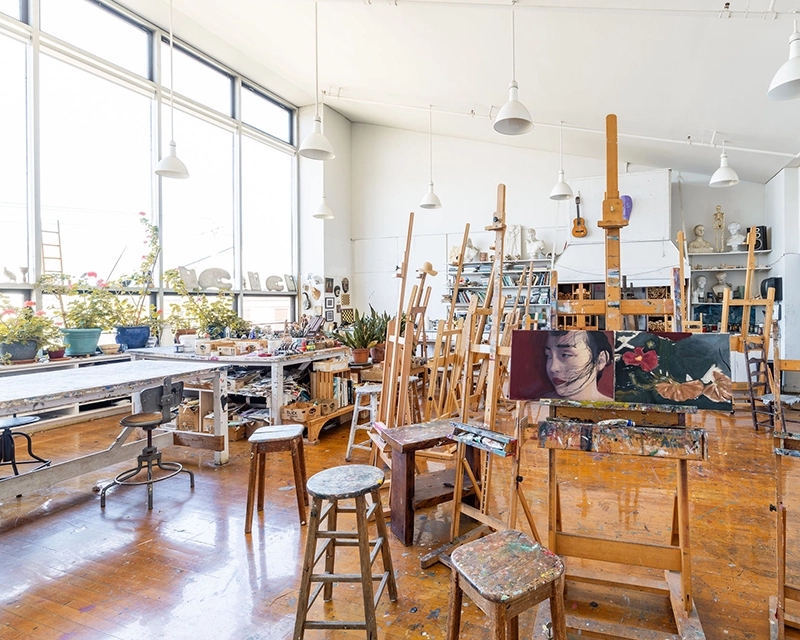 Bright art studio with large windows, wooden floors, easels, stools, and art supplies. A portrait painting of a woman is on one easel. Plants and shelves with materials line the walls.