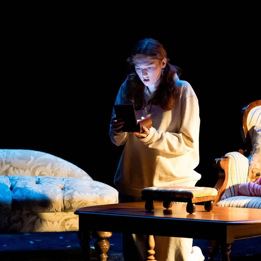 A young woman in casual clothes stands on stage, illuminated by the light from an open box she holds, with vintage furniture in the dim background.
