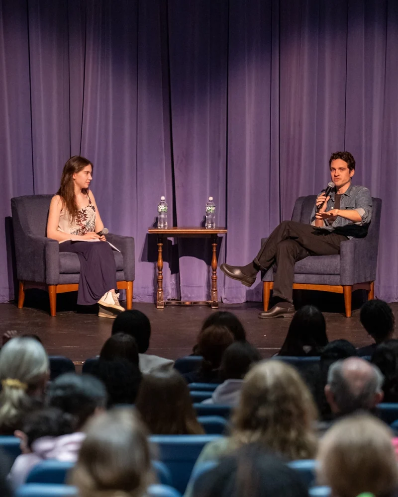 Two people sit on stage in armchairs, holding microphones, engaged in conversation. A small table with two water bottles is between them. An audience watches in a theater with purple curtains as the backdrop.
