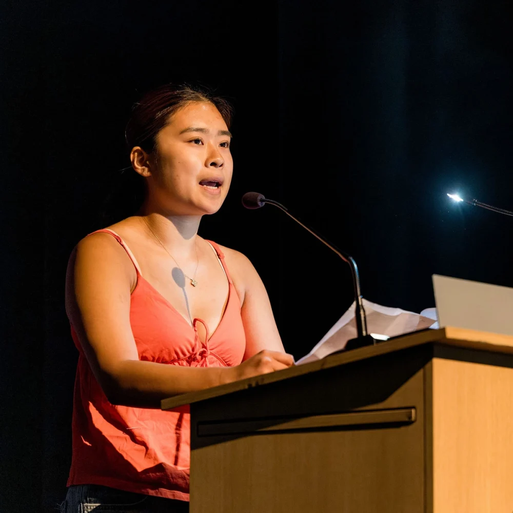 A woman in a coral tank top speaks at a podium with a microphone, holding papers under a spotlight against a dark background.