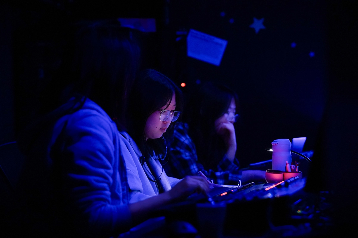 Three people work at illuminated control boards in a dark room, focused on screens and equipment, with blue and purple lighting creating a technical, behind-the-scenes atmosphere.