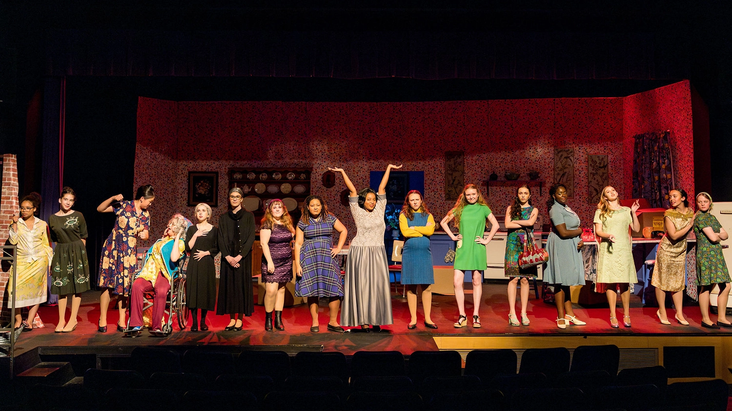 A group of people in colorful dresses stand on stage in a dramatic pose, with a theater set featuring patterned wallpaper and furniture in the background.