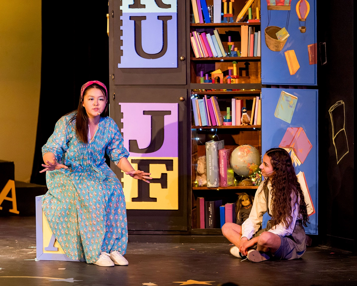 Two people perform on stage; one in a blue dress sits on a block with letters, gesturing, while the other, in a school uniform, sits cross-legged on the floor near colorful bookshelves and props.