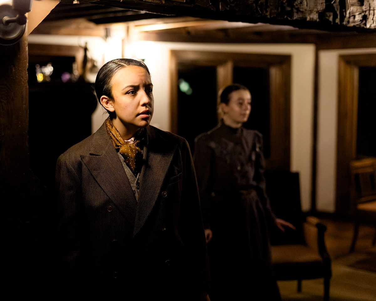 Two women in old-fashioned clothing stand in a dimly lit room with wooden beams and large windows. One woman is in the foreground looking worried; the other is in the background, partially out of focus.