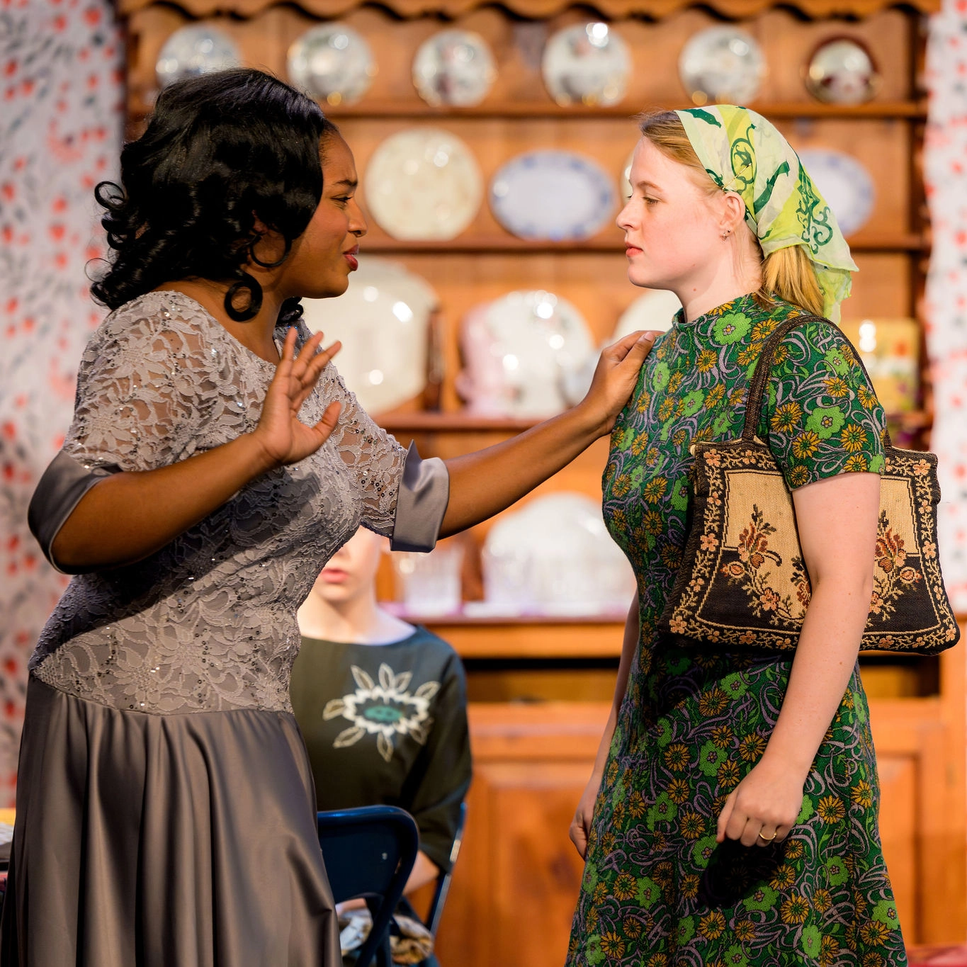 Two women are standing indoors, one wearing a gray dress with lace, gesturing with her hand, while the other, in a green floral dress and headscarf, holds a bag. They appear to be having a serious conversation.